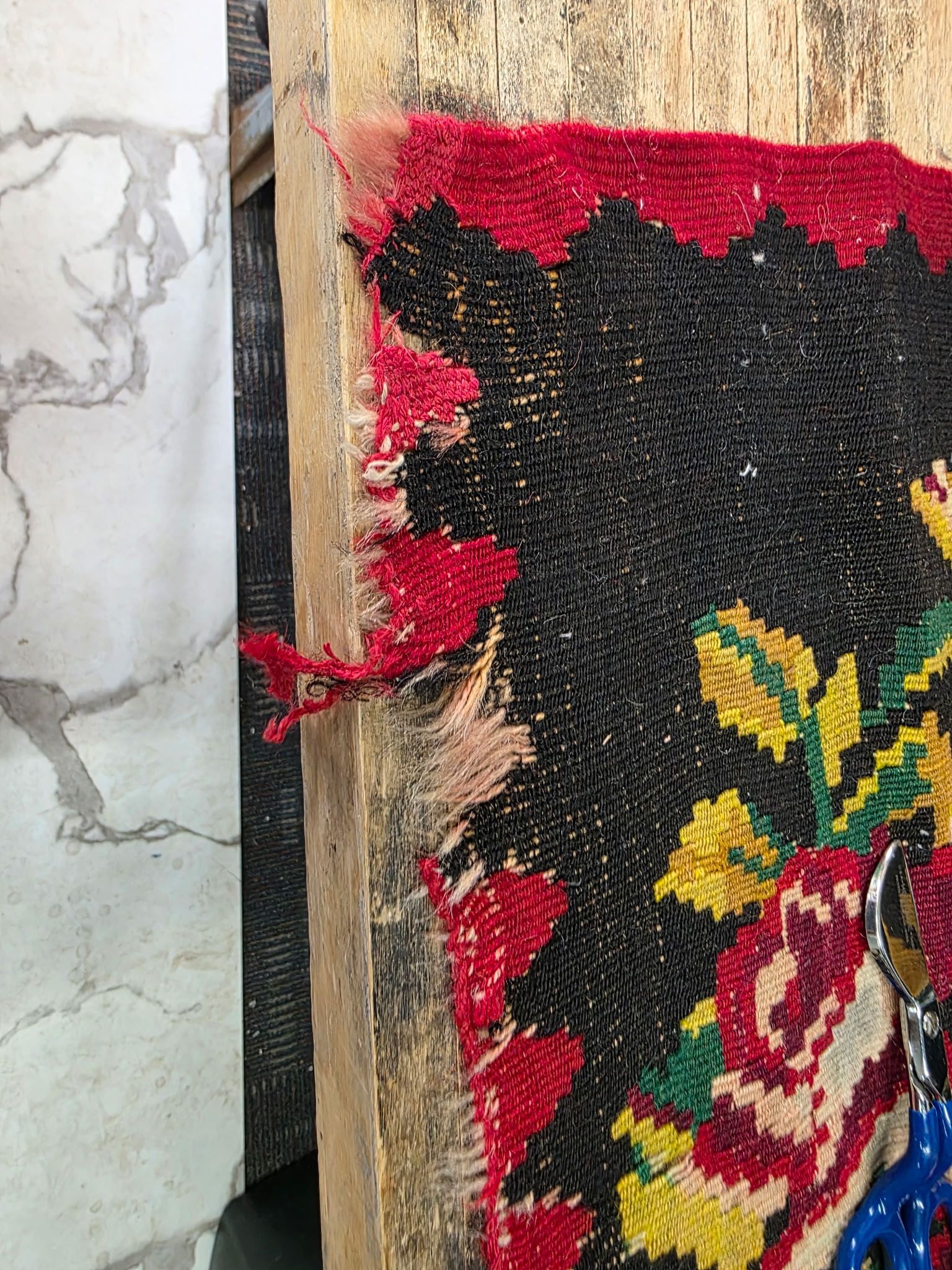 Close-up of a worn floral tapestry with red, black, yellow, and green pattern beside a cracked wall