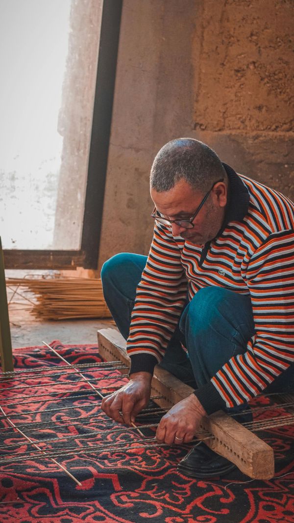 Man kneels on a patterned rug, working with tools beside a wooden board in a sunlit room.