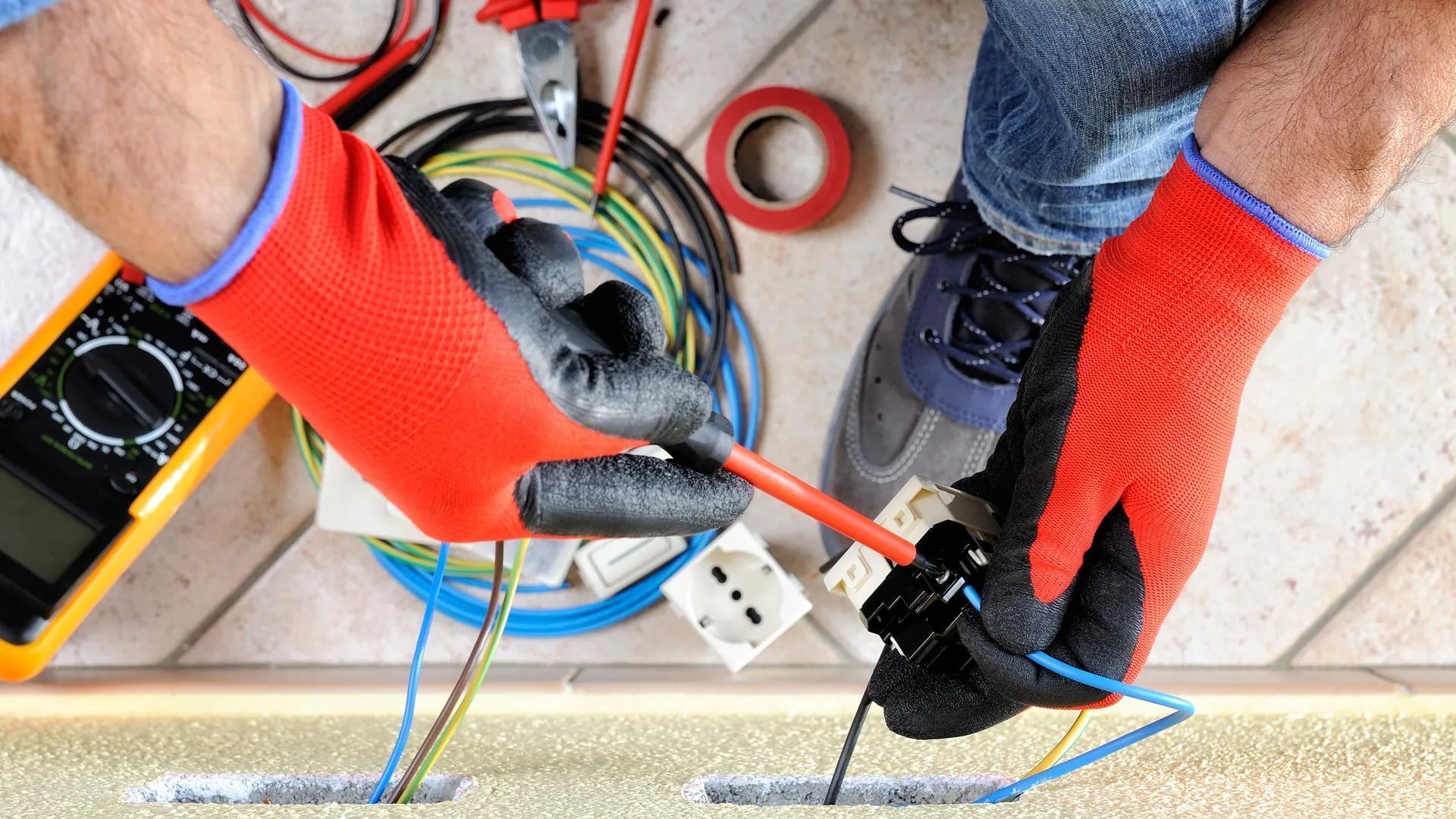 Gloved hands using a screwdriver to wire an electrical outlet box on a floor with cables nearby