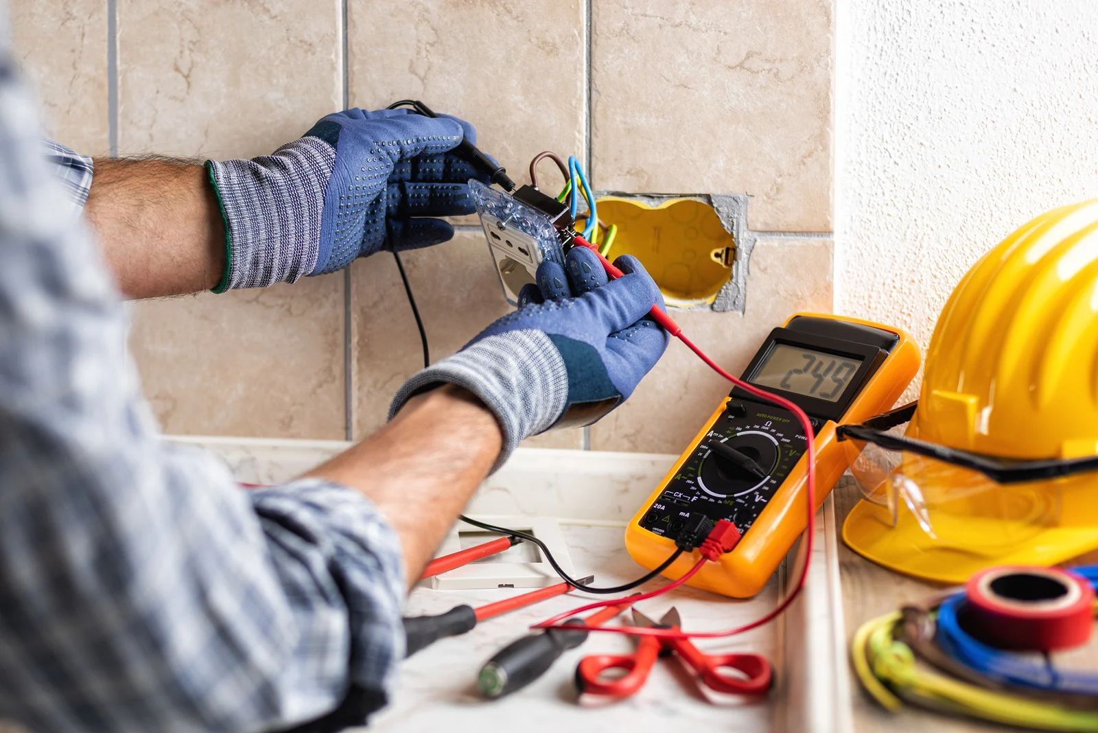 Electrician tests wires in an outlet with a multimeter, wearing gloves.