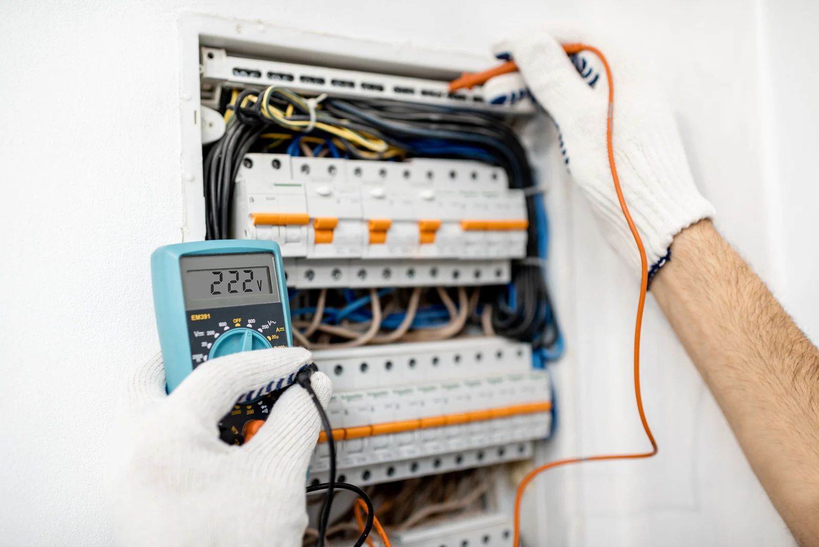 Electrician testing a fuse box with a multimeter, wearing gloves.