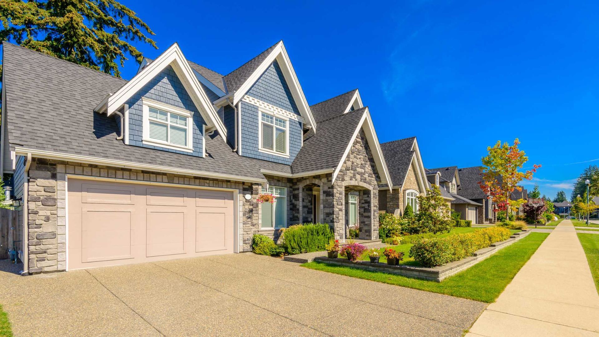 Suburban stone house with gray roof, peach garage doors, and a sunny front lawn along a sidewalk.