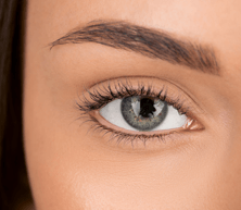 Eine Nahaufnahme eines menschlichen Auges mit grauer Iris, langen dunklen Wimpern und natürlich geformten Augenbrauen.