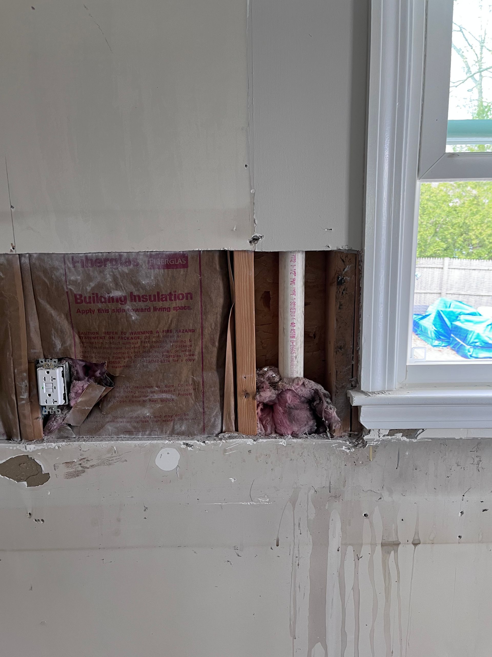 Partially opened wall reveals insulation and studs beside a white-framed window with blue tarp outside