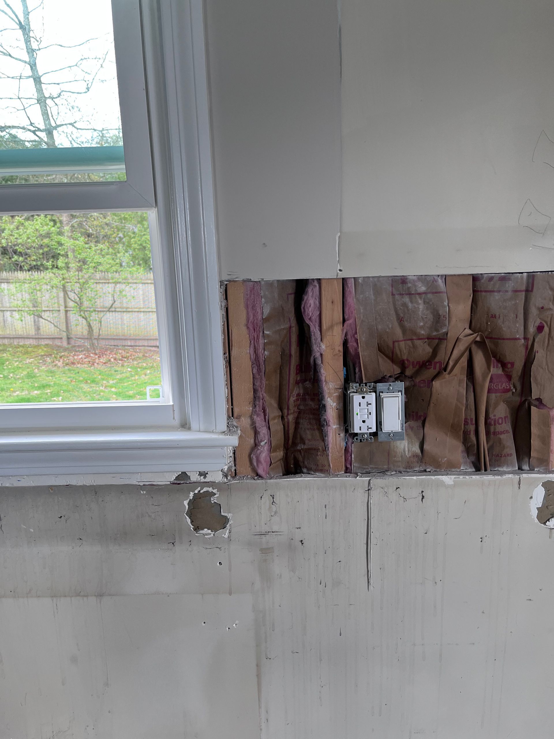 Wall section stripped to studs beside a window, exposing electrical wiring and outlets.