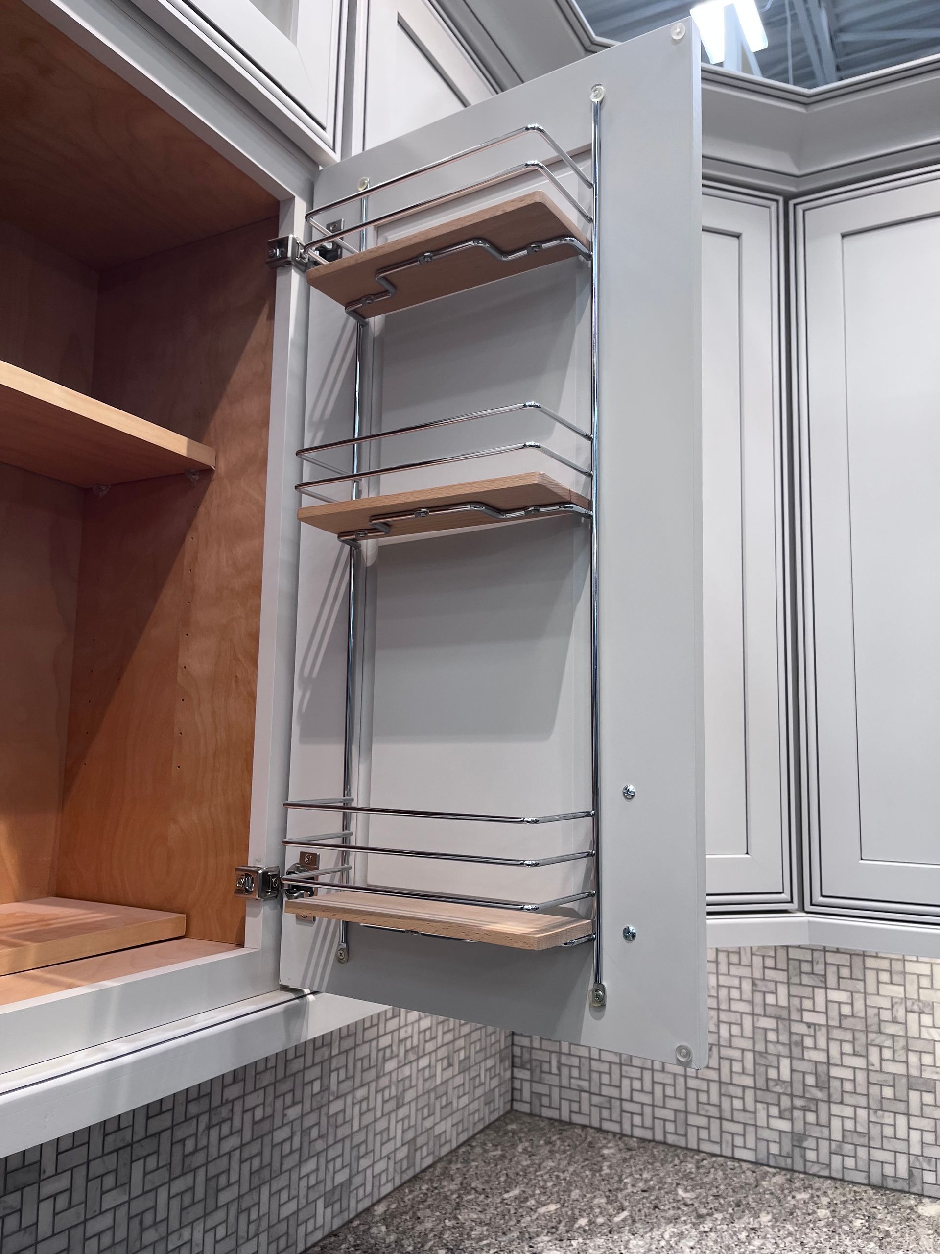 Pull-out wire pantry shelves inside an open kitchen cabinet with white doors and a tiled backsplash.
