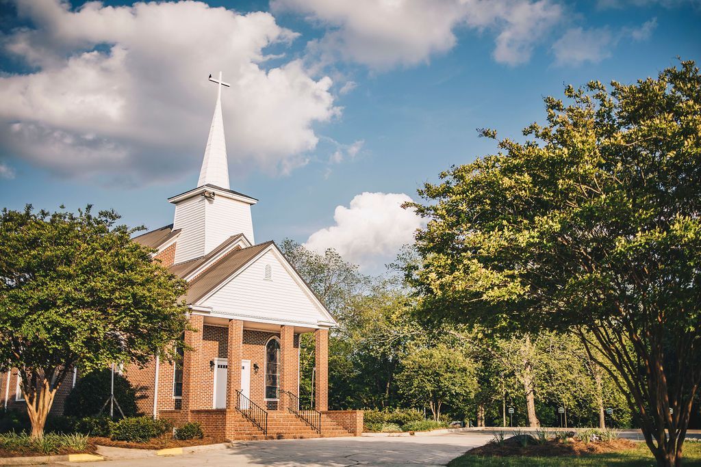 A small church with a white steeple is surrounded by trees on a sunny day.