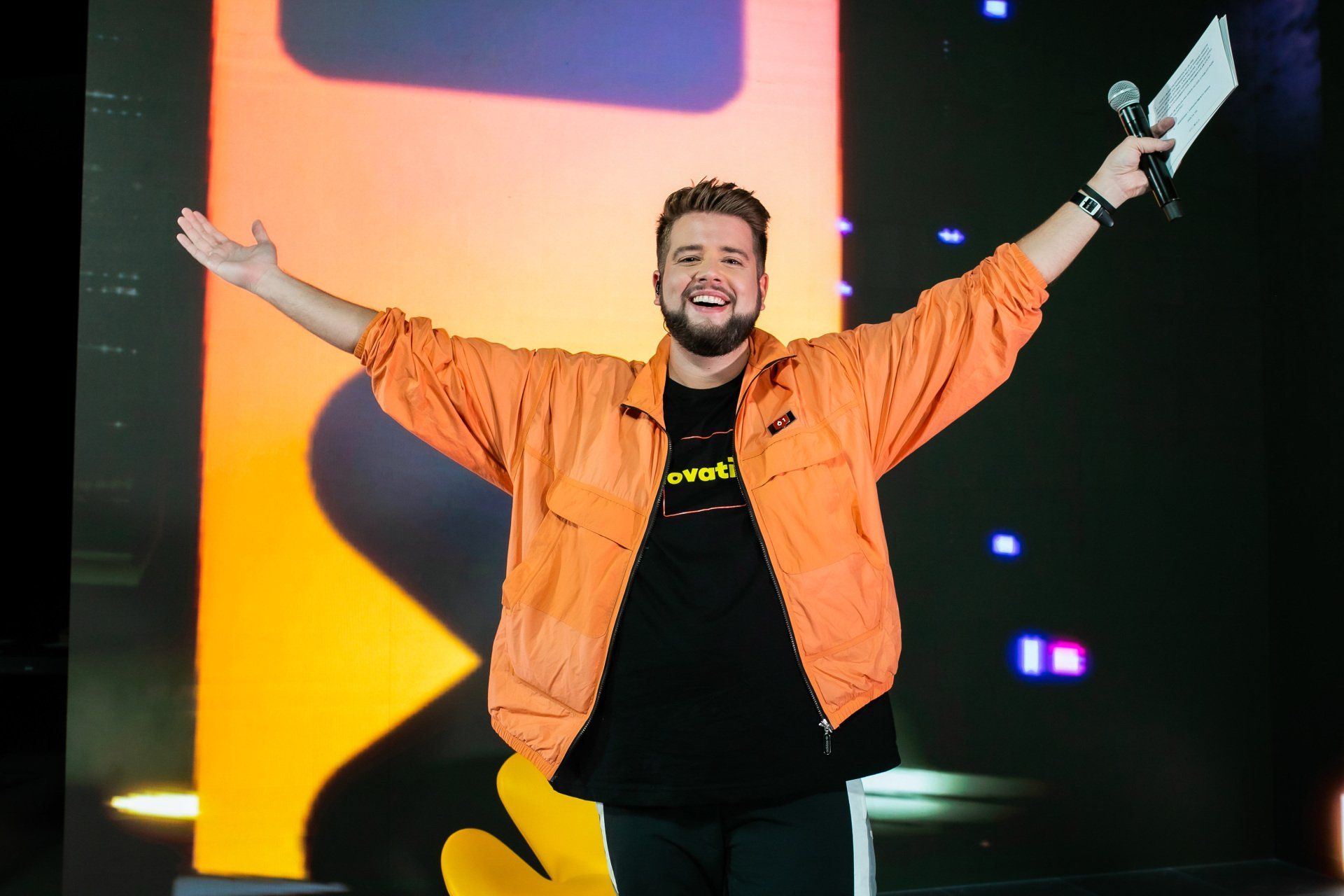 A man in an orange jacket is holding a microphone in front of a large screen.