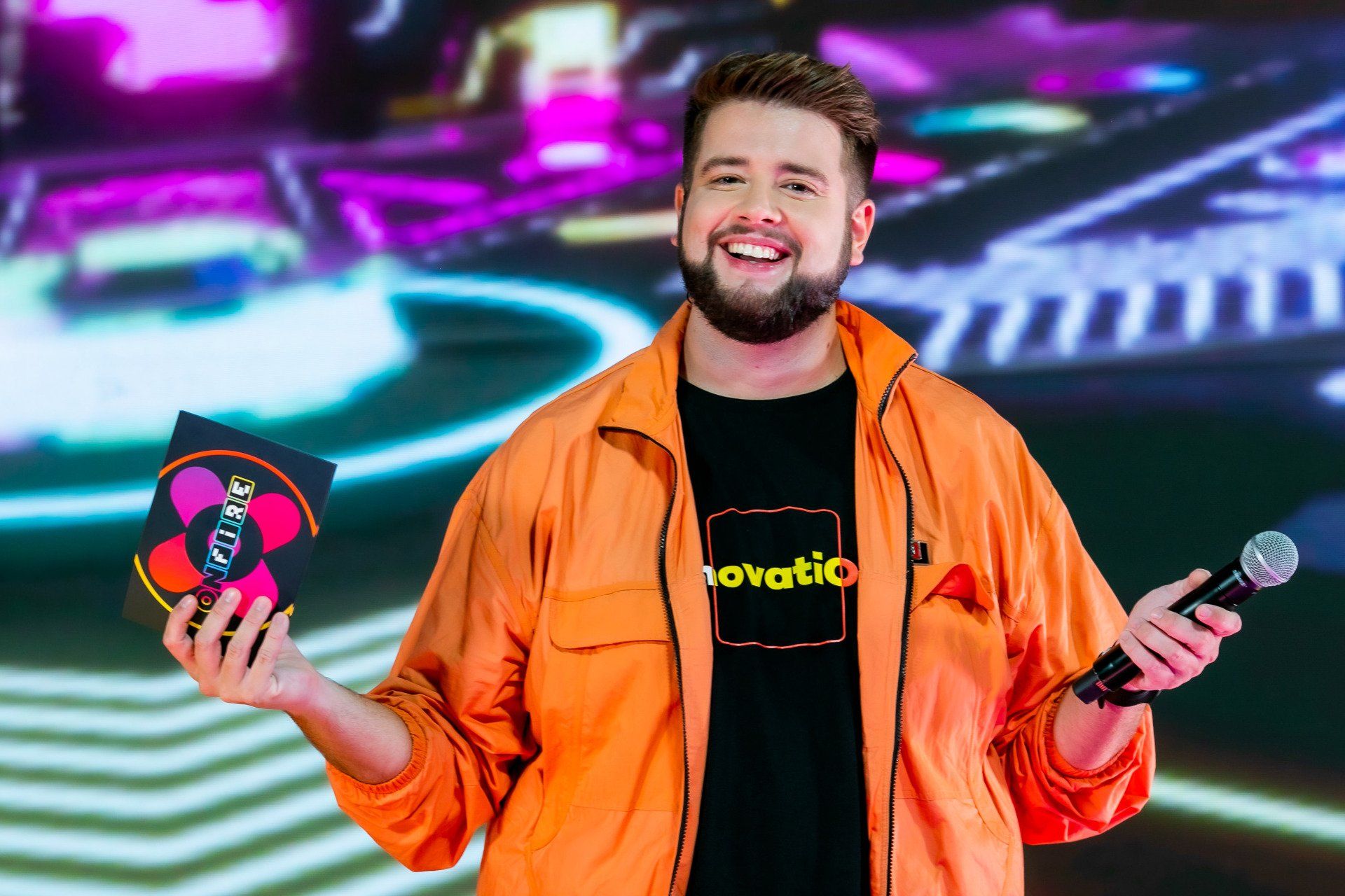 A man in an orange jacket is holding a microphone and a cd.