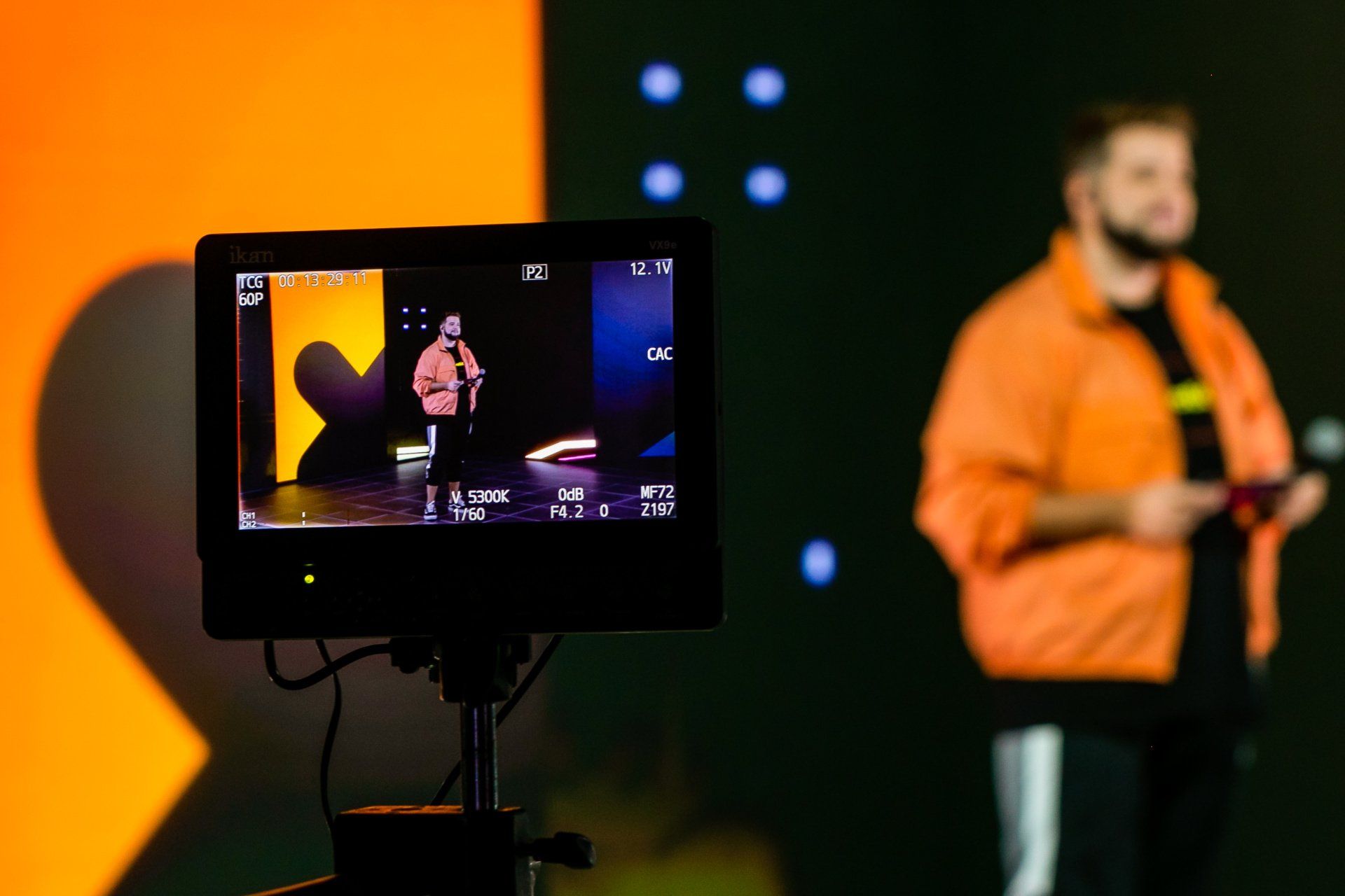 A man in an orange jacket is standing in front of a camera.