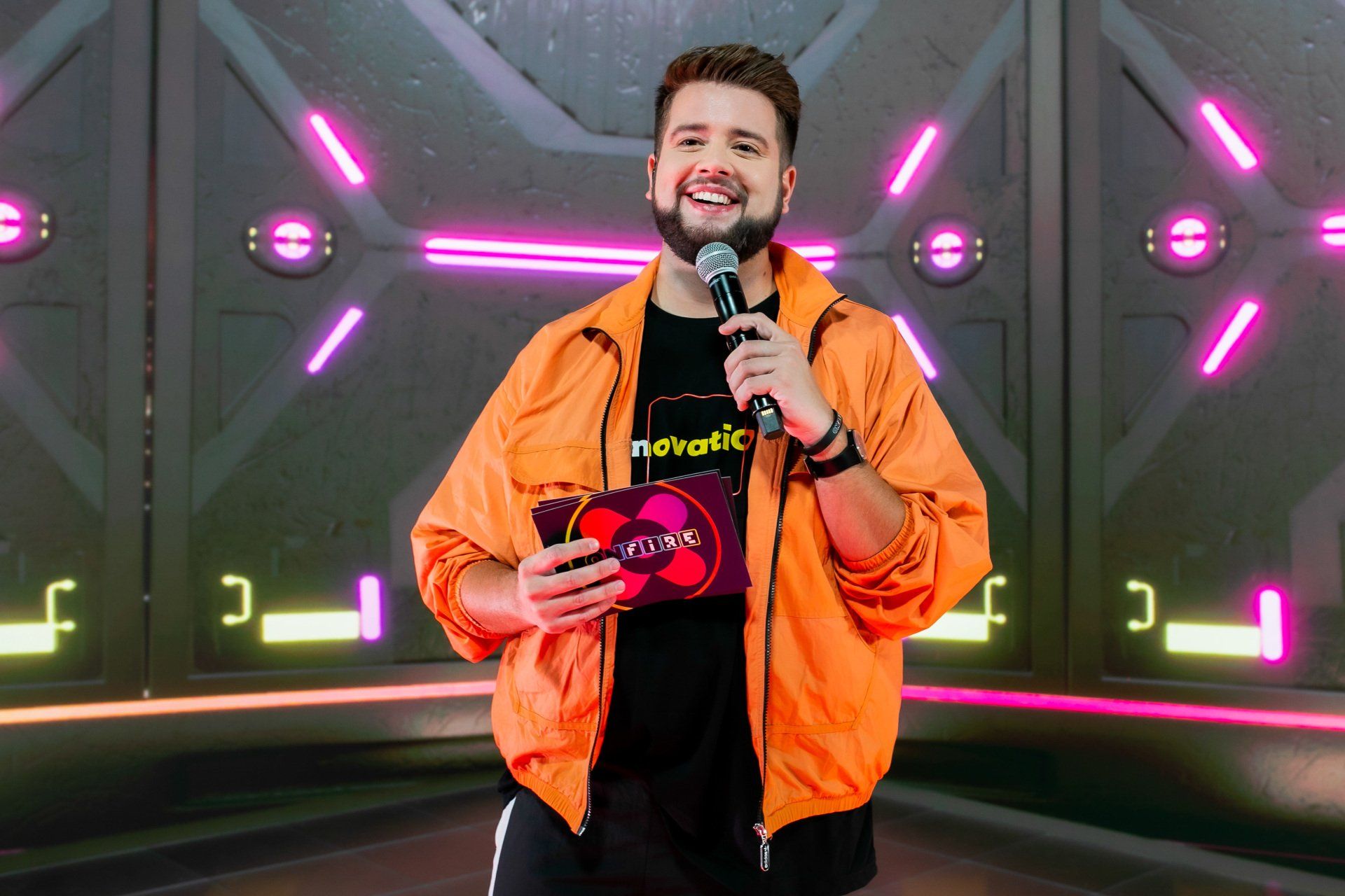 A man in an orange jacket is holding a microphone in front of a neon wall.