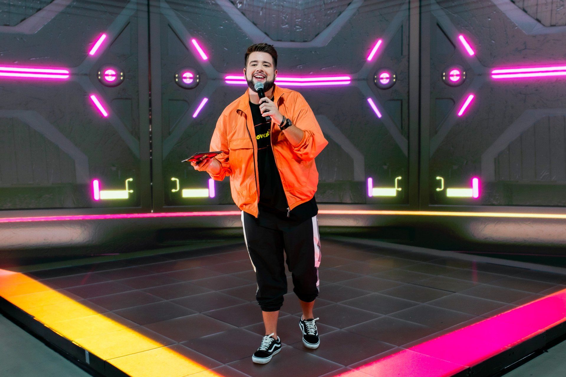 A man in an orange jacket is standing on a stage holding a microphone.