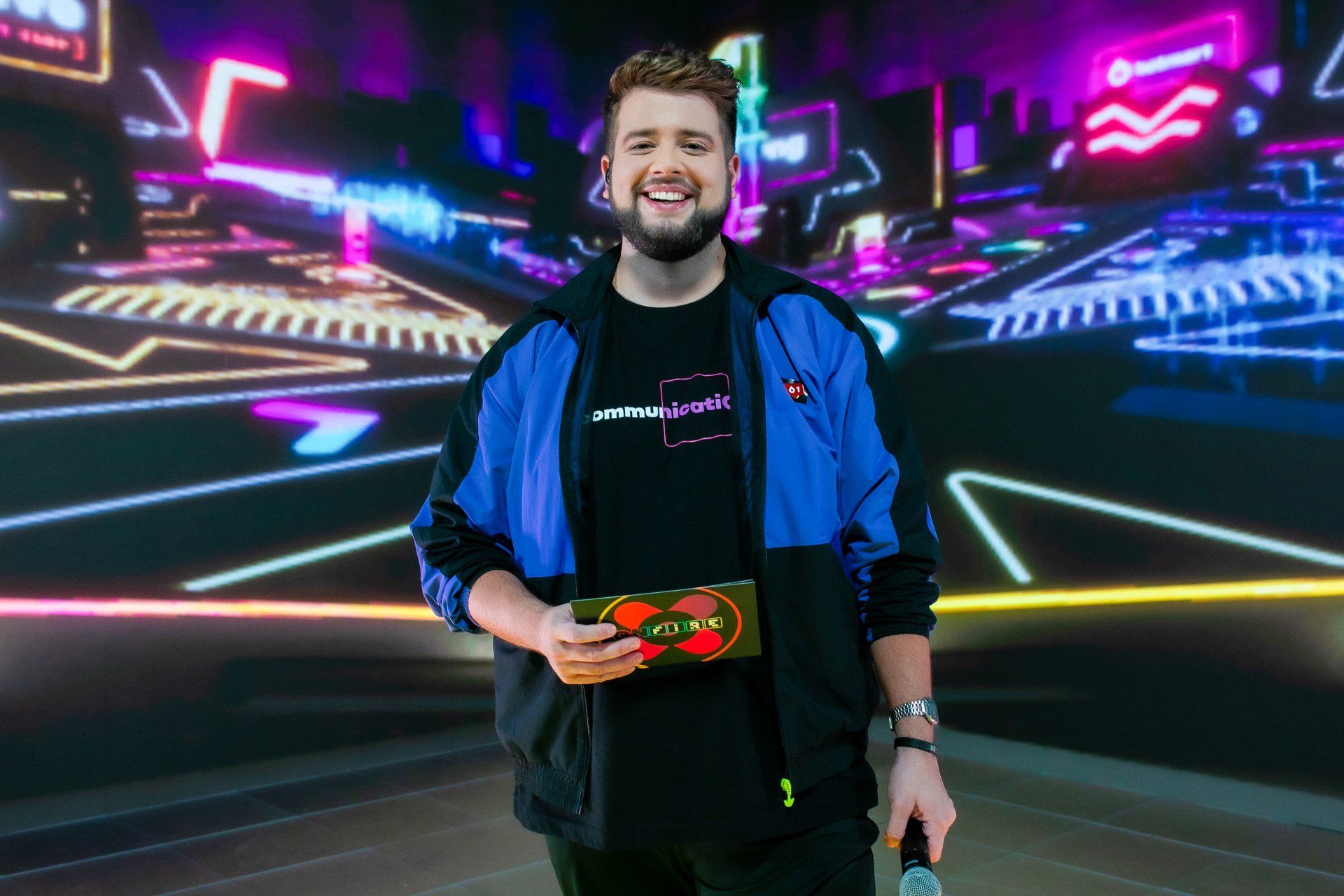 A man in a blue jacket is holding a microphone in front of a neon background.