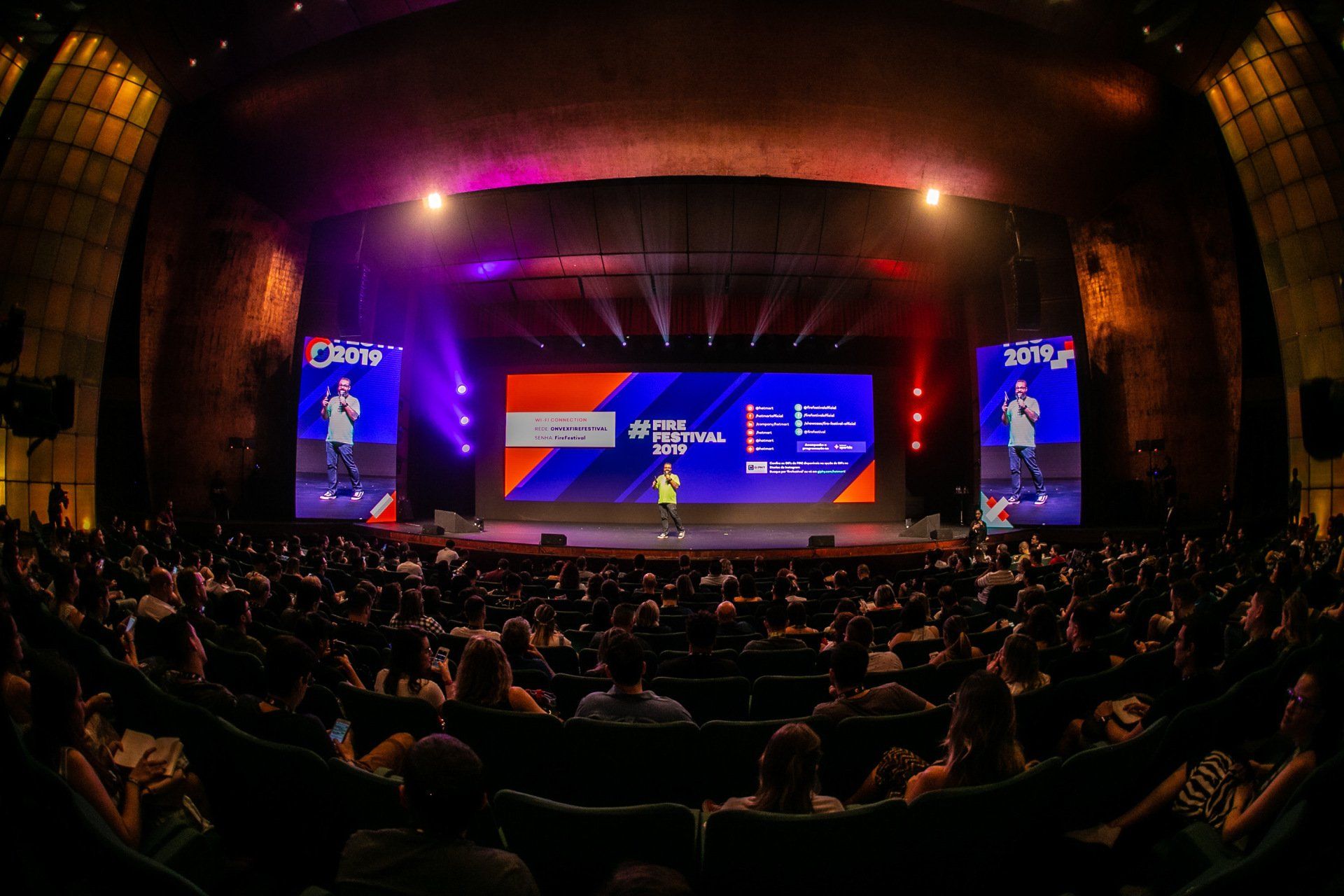 A large crowd of people are sitting in a theater watching a presentation on a stage.