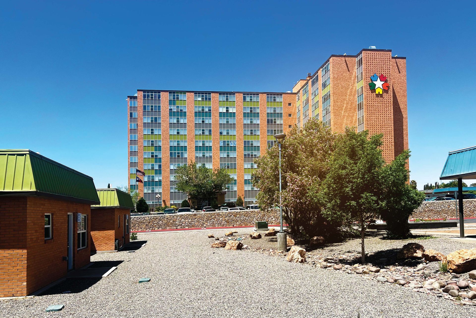 A tall, orange-brick building with a colorful logo stands behind a gravel yard with small, green-roofed brick structures.