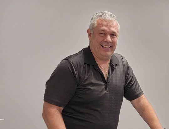 A man in a black shirt is smiling and standing in front of a white wall.