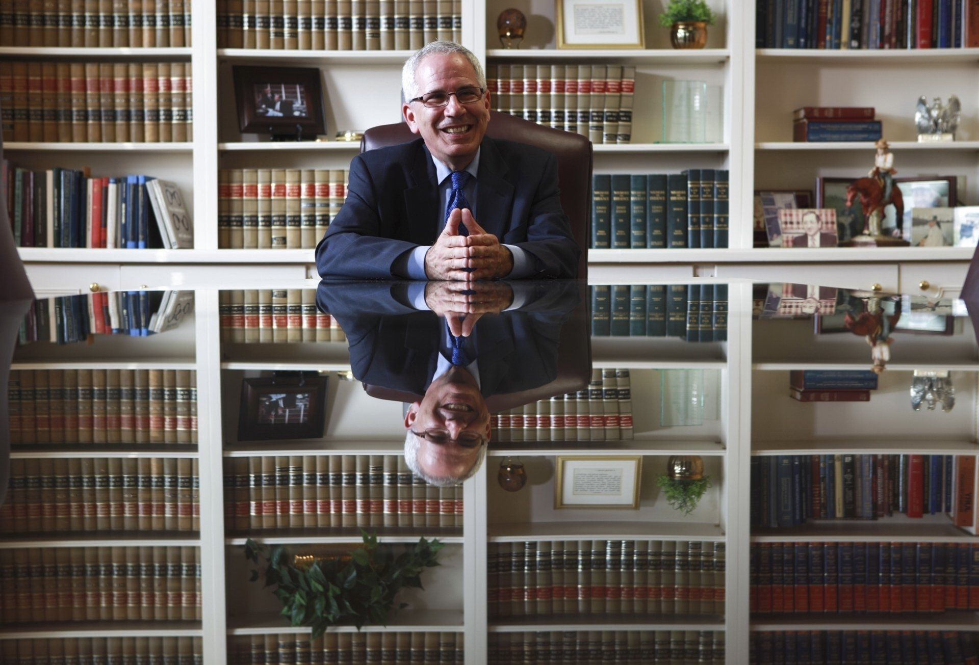 A man in a suit sits at a desk in front of a bookshelf