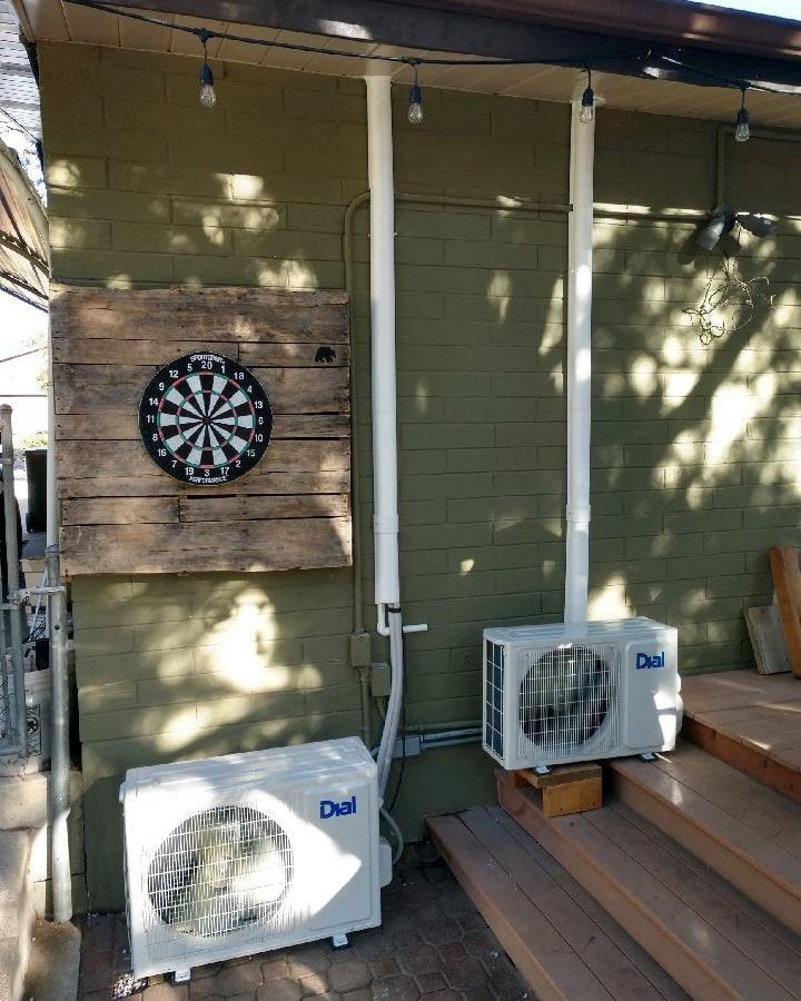 Dartboard on a wooden plank, next to two outdoor air conditioning units on a green building.