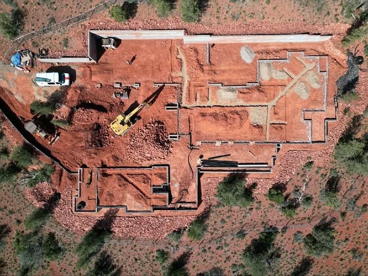Overhead view of a house foundation under construction on reddish soil; excavator, materials, and equipment visible.