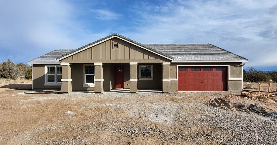 Tan one-story house with red garage door, on a gravel lot under a blue sky.