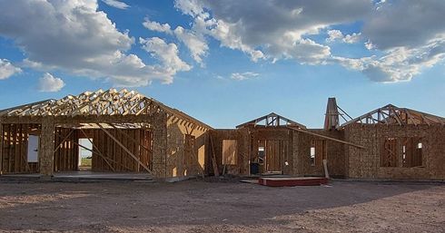 Wood-framed house under construction on a dirt lot, against a blue sky with fluffy clouds.