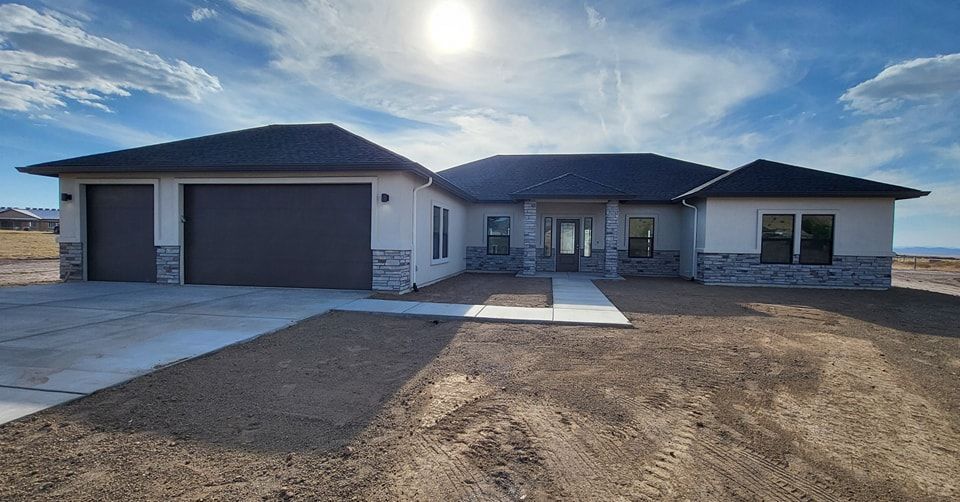 A single-story house with a two-car garage under a bright sun. Gray stucco with stone accents.