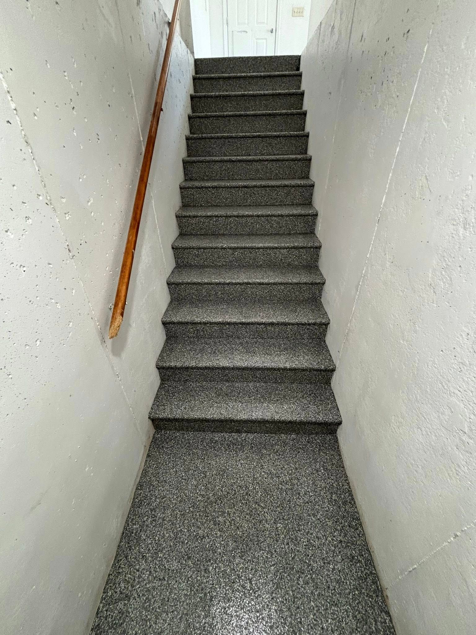 Staircase with gray speckled steps and a wooden handrail, leading upwards between gray walls.