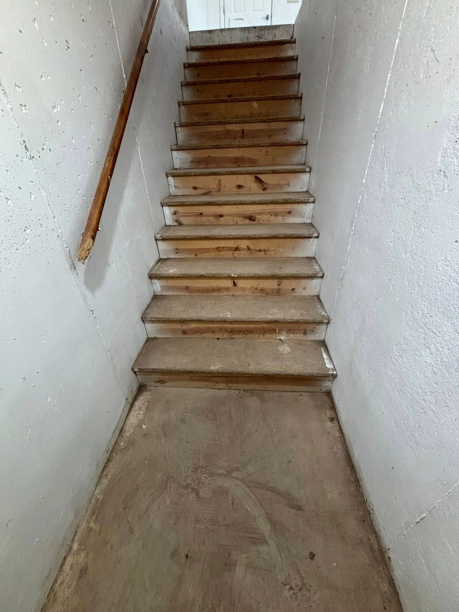 Wooden staircase descending into a dingy, light-colored room. Handrail on the left.