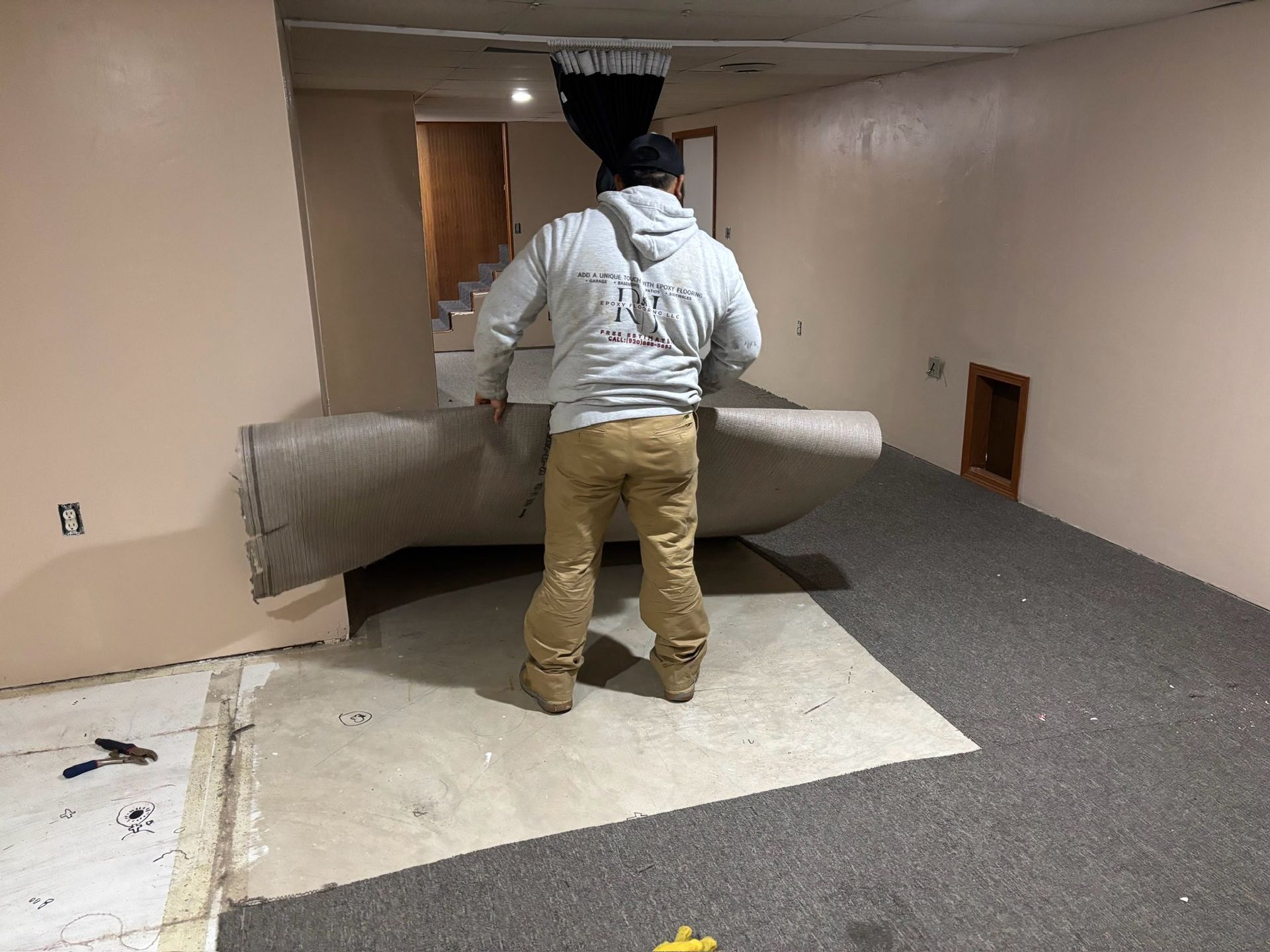 Person rolls out new carpet in a room with tan walls and concrete flooring.