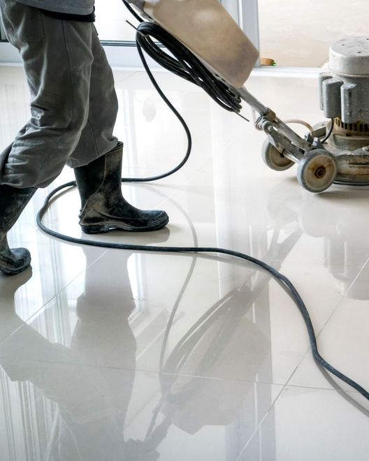 Person in rubber boots polishing a shiny, white tiled floor with a floor buffer.