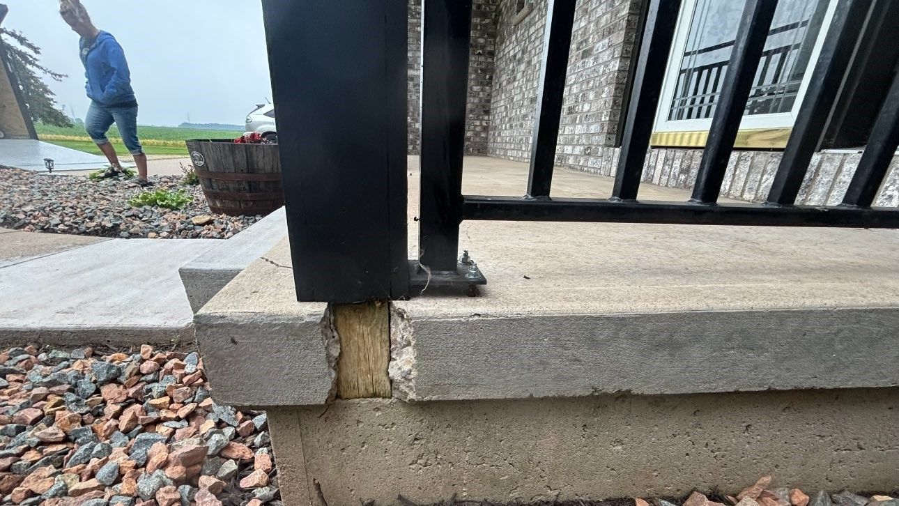 Close-up of a concrete porch with a black railing, wooden support, and gravel. A person in a blue jacket is in the background.