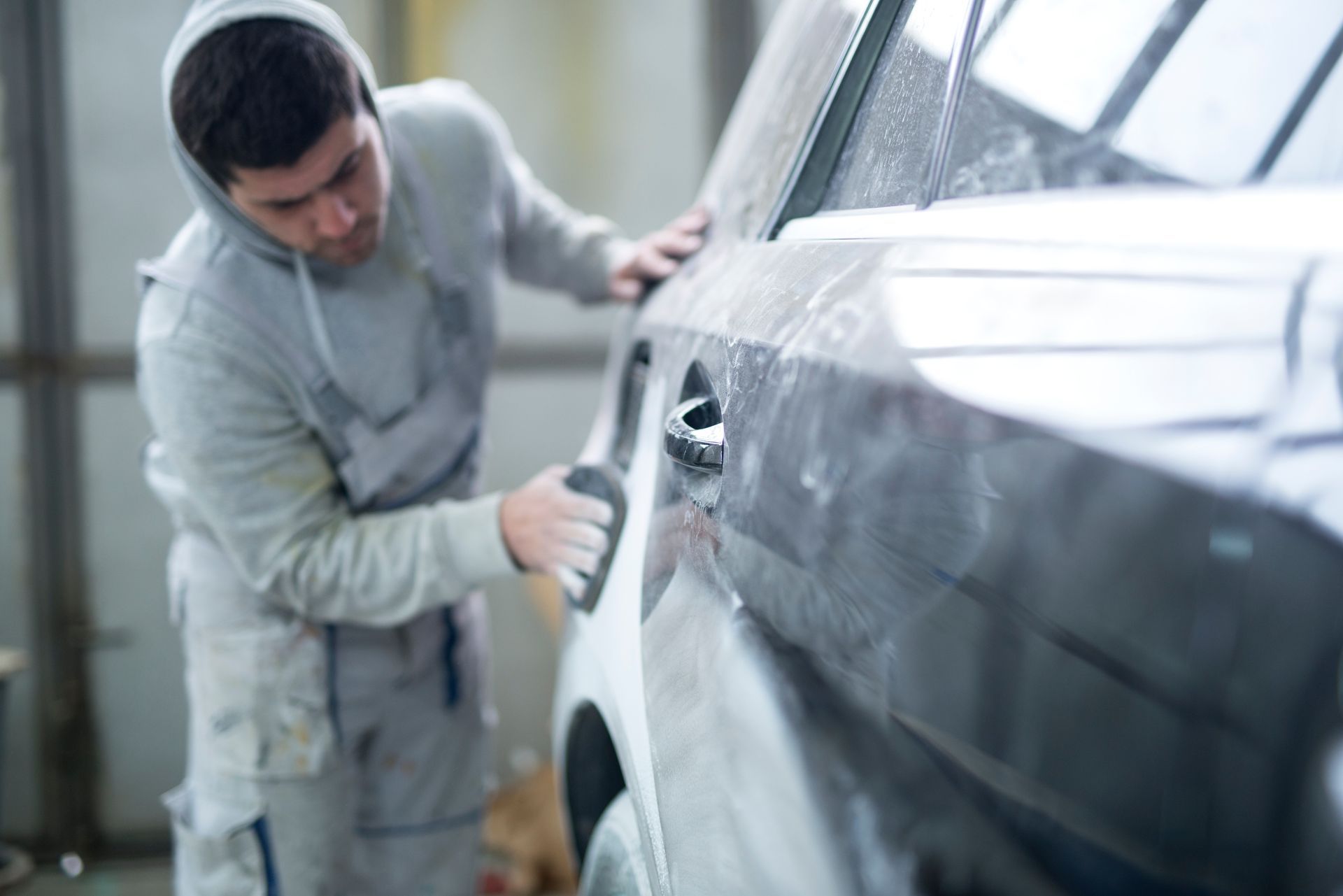 Technician in gray hooded coveralls sanding a car door panel during collision repair in a shop bay.