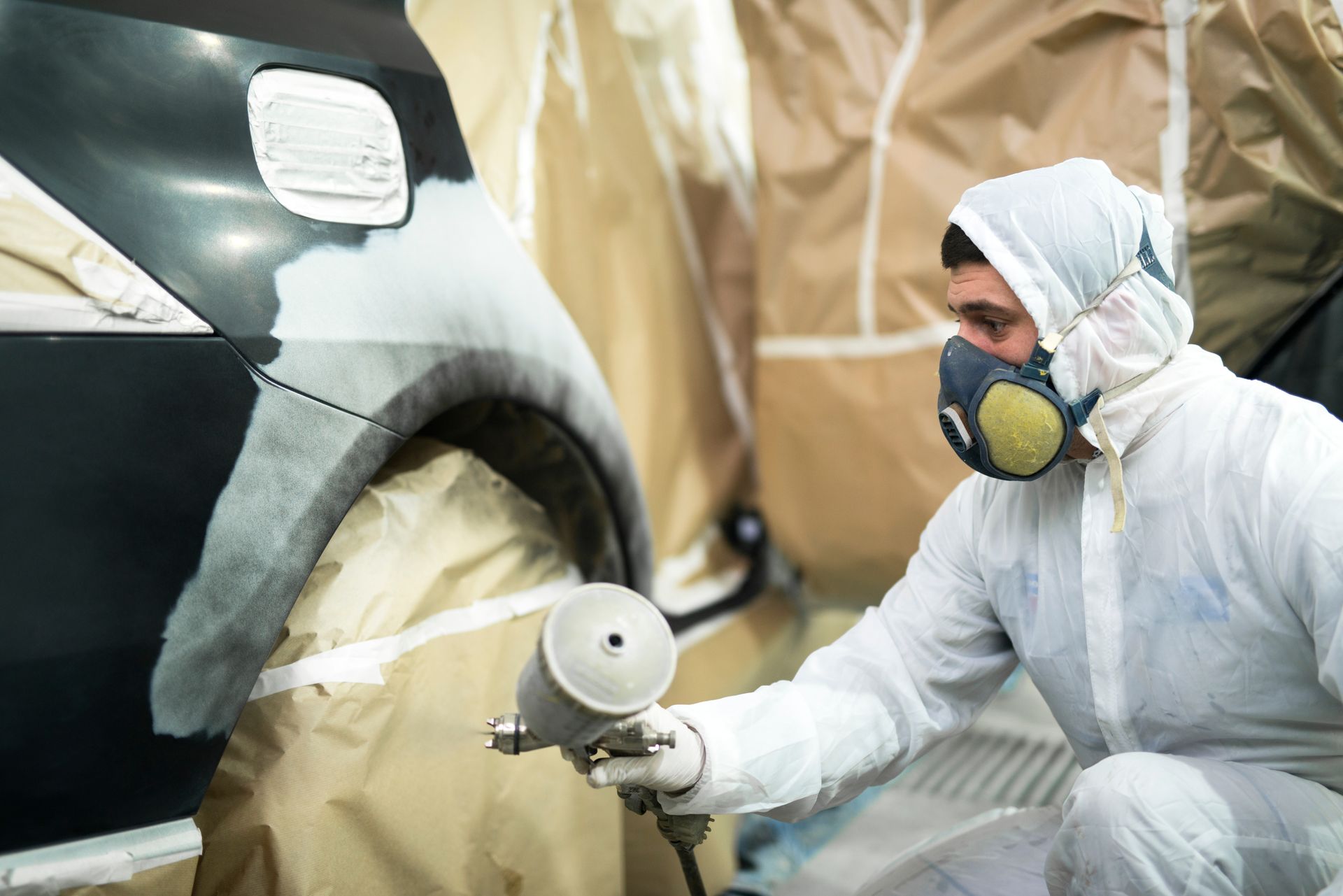 Worker in protective suit spray-painting a car body in an auto repair booth.