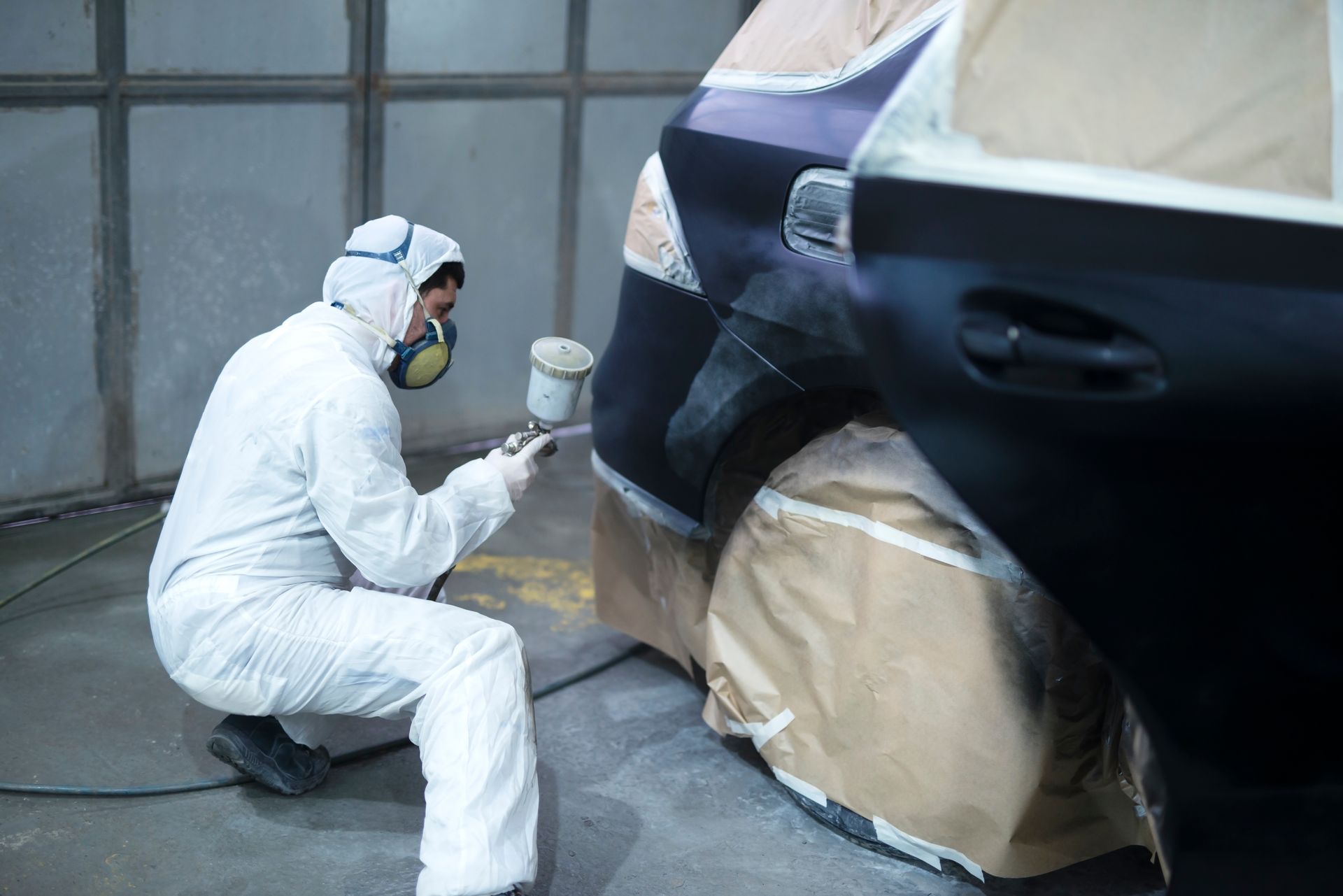 Worker in white coveralls spray-painting a car panel in a masked auto body shop.