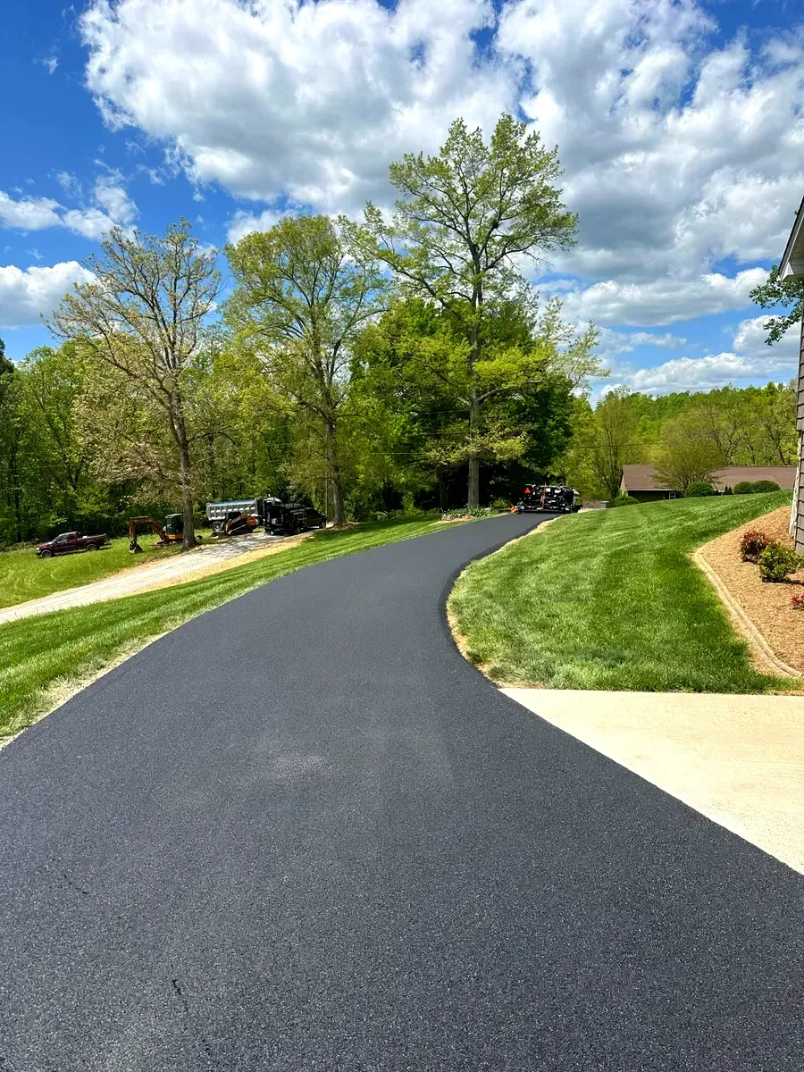 Newly paved asphalt driveway curves through a green yard, trees in the background, under a blue sky with clouds.