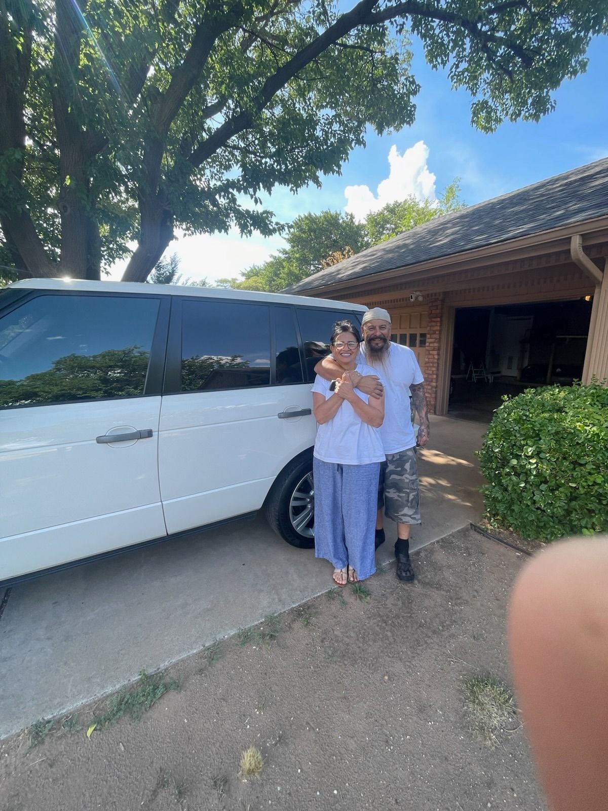 Person and child standing beside a white van on a driveway under trees, with a house in the background