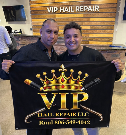 Two men holding a VIP Hail Repair LLC banner in a repair shop with a VIP Hail Repair sign behind them