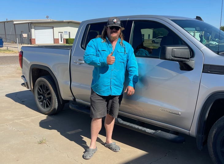 Person in blue jacket and hat stands beside a silver pickup truck outdoors, giving a thumbs-up.