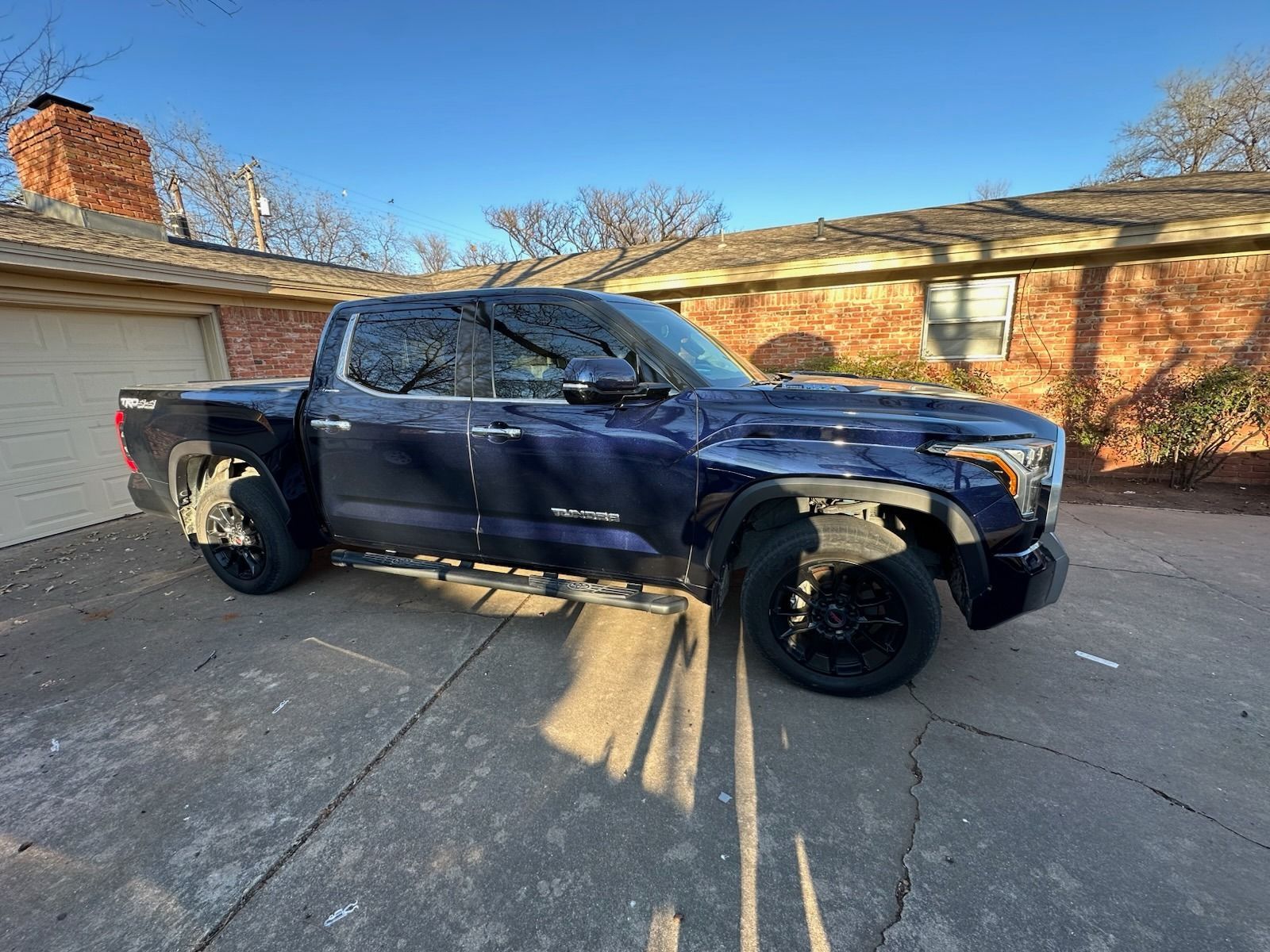 Blue pickup truck parked in a driveway beside a garage and brick house, with front wheel damage
