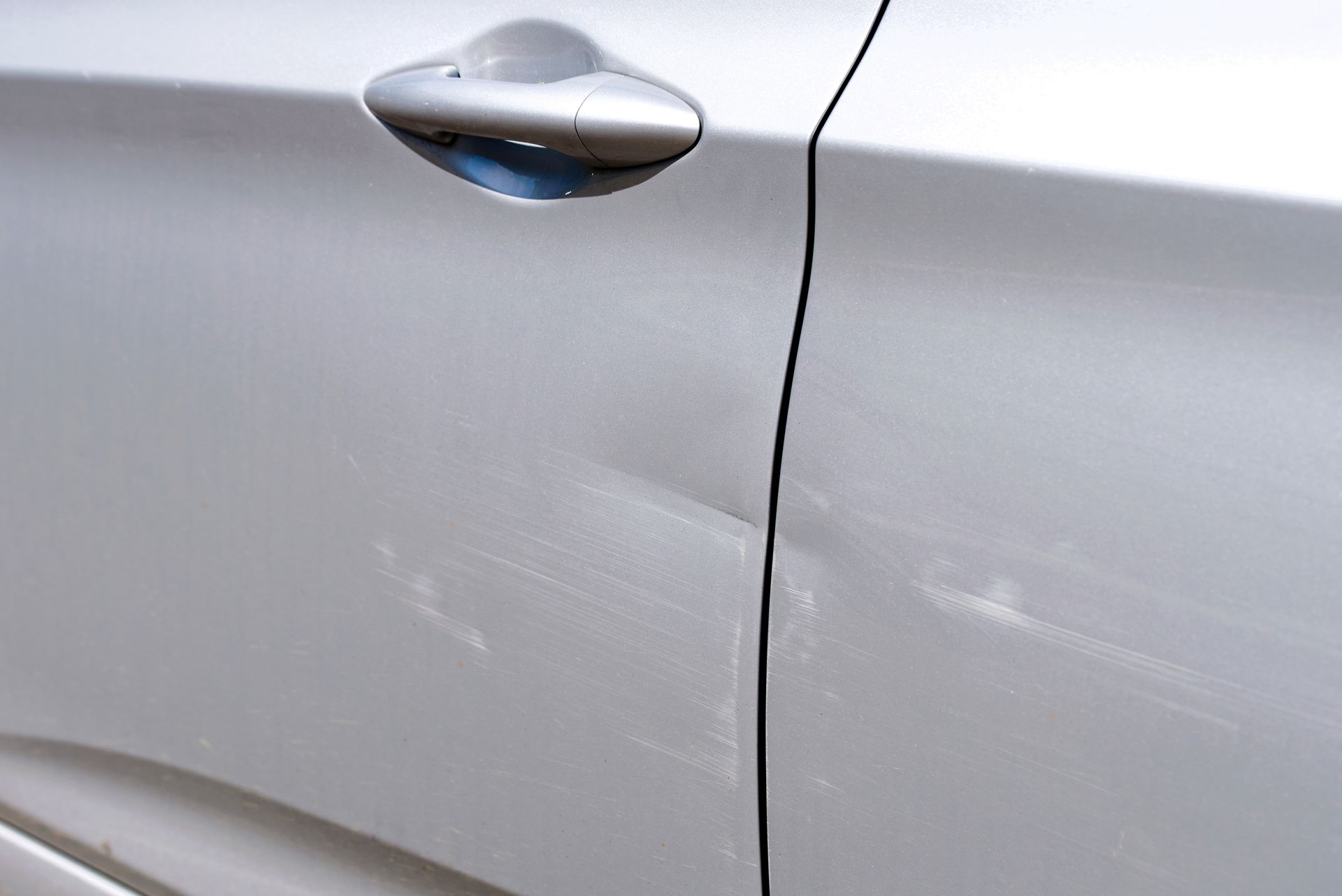 Close-up of a silver car door with scratches near the handle and panel seam