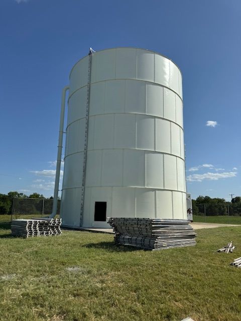 White cylindrical water tower against a bright blue sky. A metal ladder runs up the side; concrete blocks are stacked nearby.