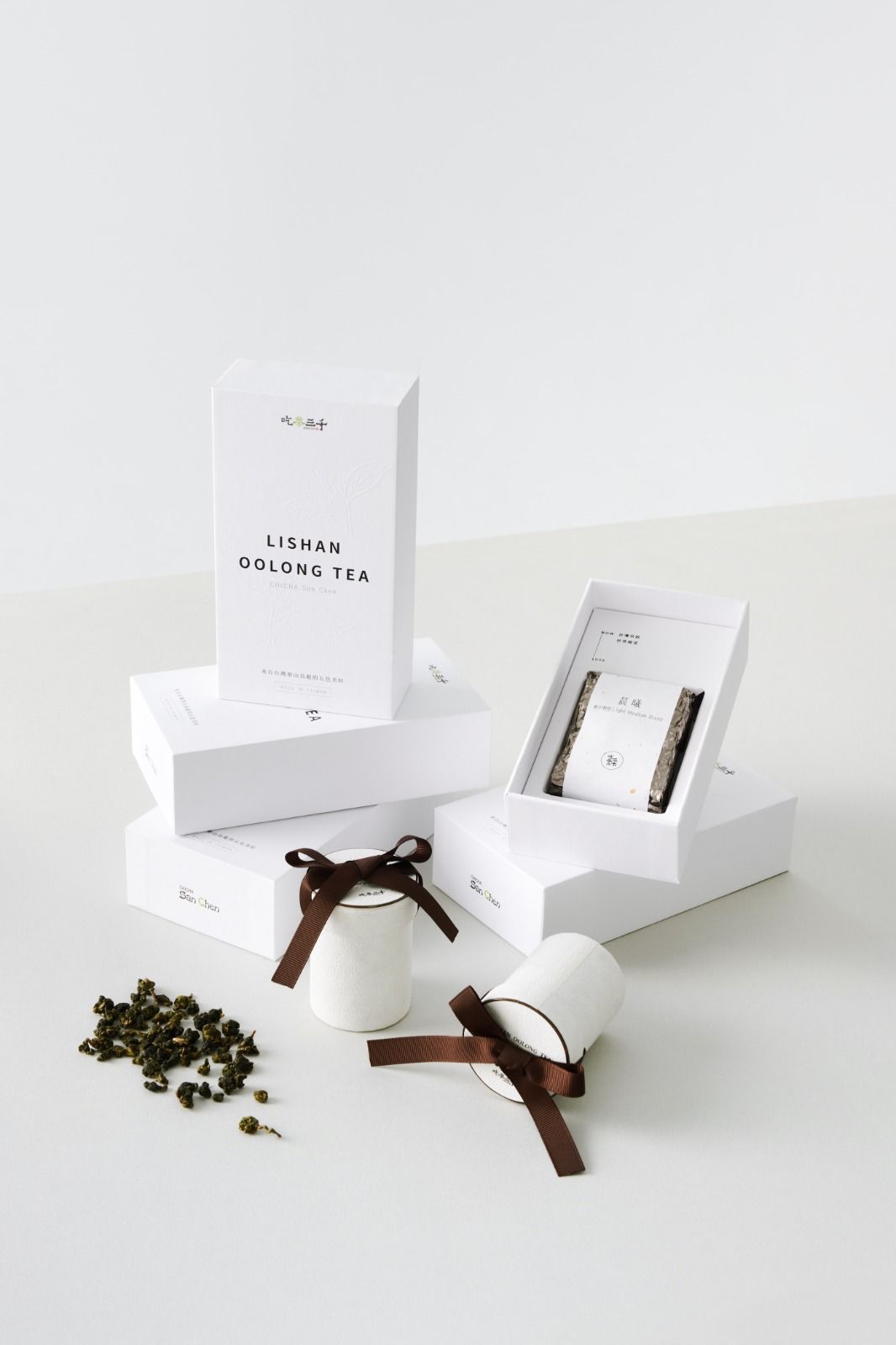 White skincare boxes with brown ribbon bows, loose tea leaves, and a white scoop on a clean tabletop