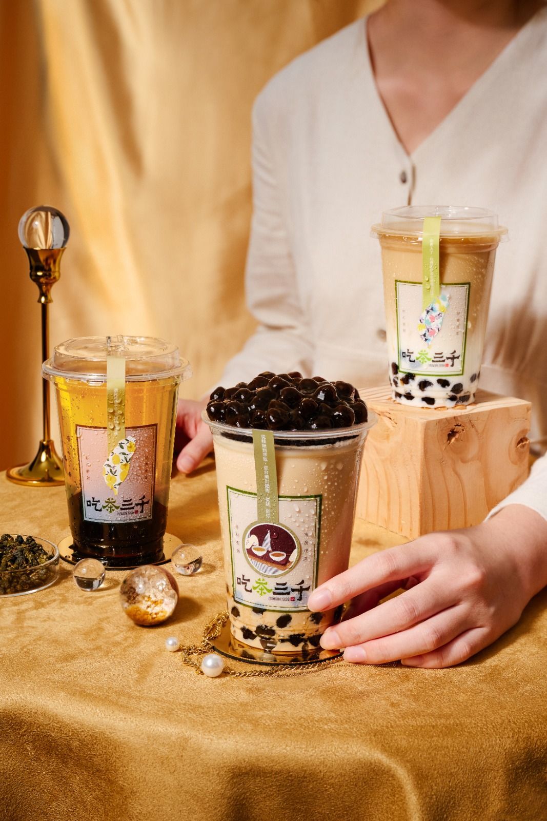 Three boba drinks in clear cups on a gold cloth, topped with whipped cream and pearls, with a person behind them