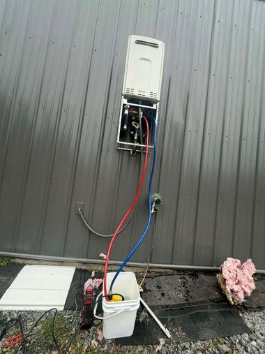 A tankless water heater mounted on a gray wall, connected by red and blue hoses to a white bucket containing a small pump.