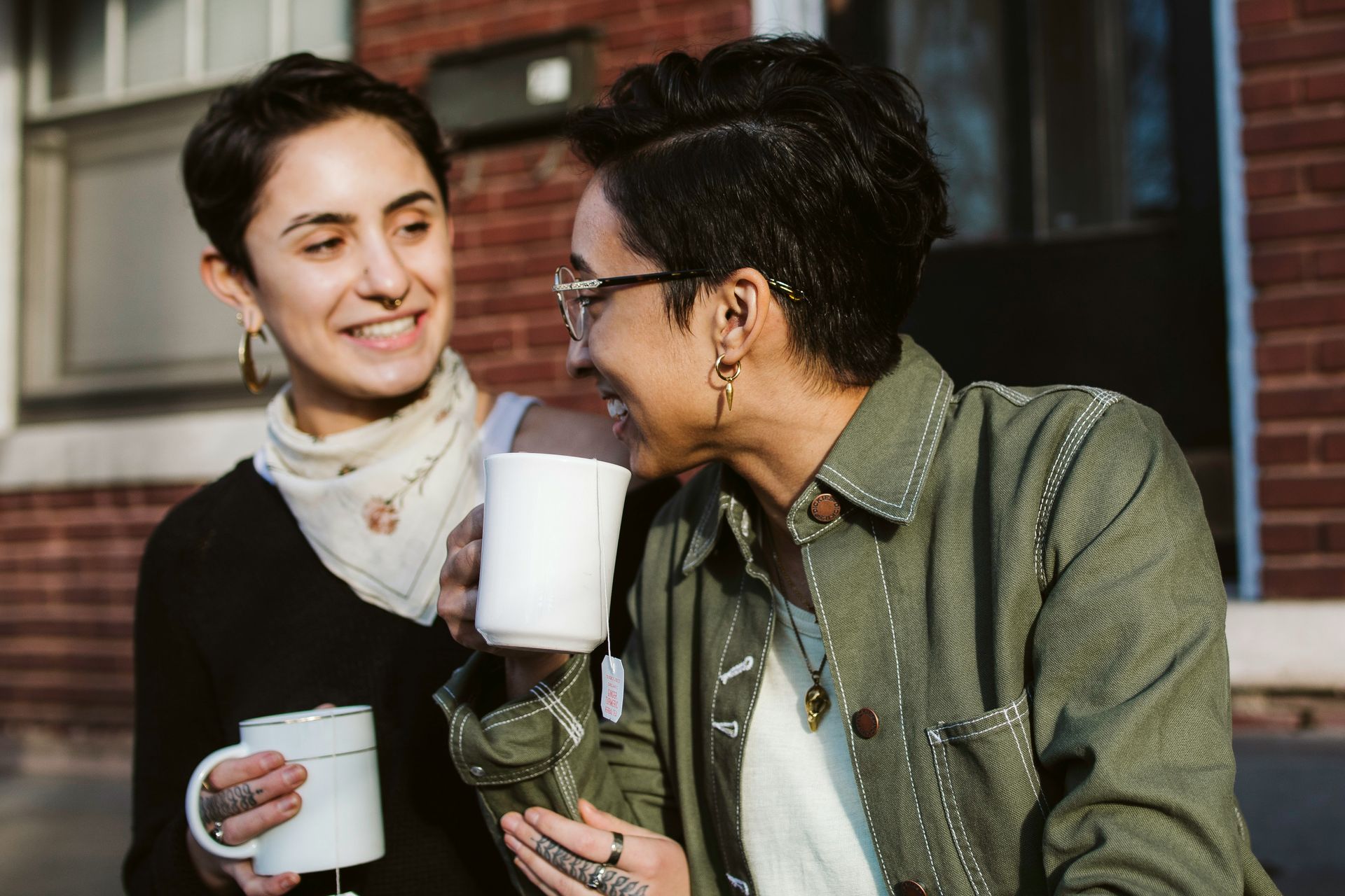 Two people with short hair laughing while holding white mugs, sitting outside in front of a red brick building.