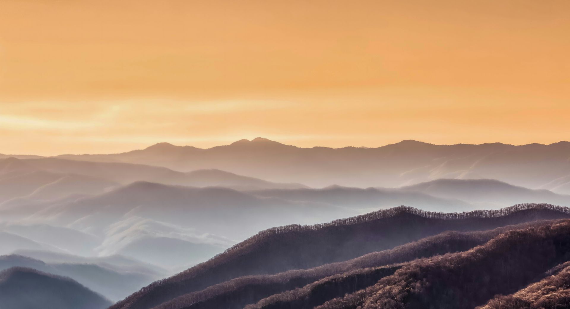 Layers of blue-toned mountain ridges fade into the distance under a warm, golden, hazy sunset sky.