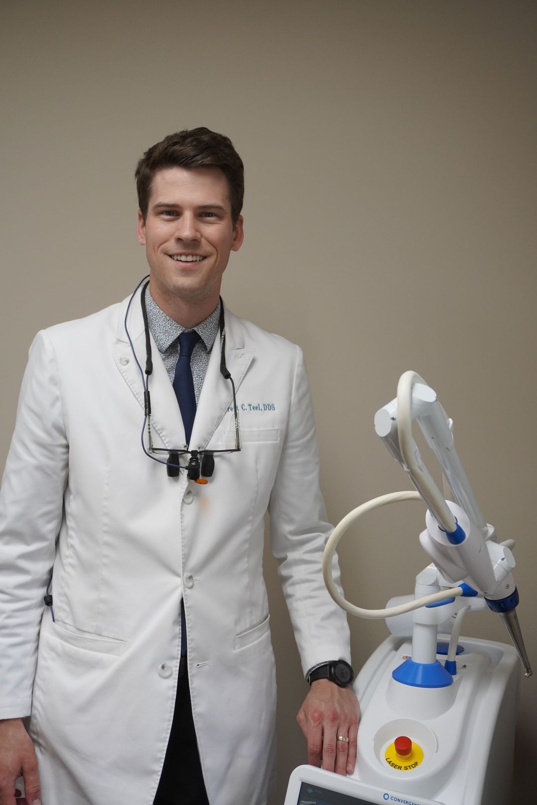 Smiling doctor in white coat, leaning on a medical device. Beige wall.