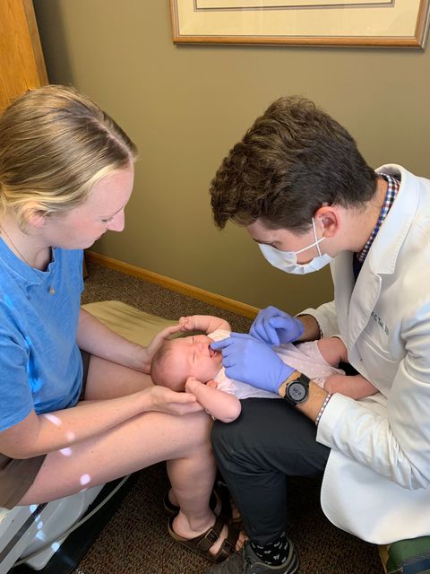 A doctor examining a baby held by a person in a blue shirt.