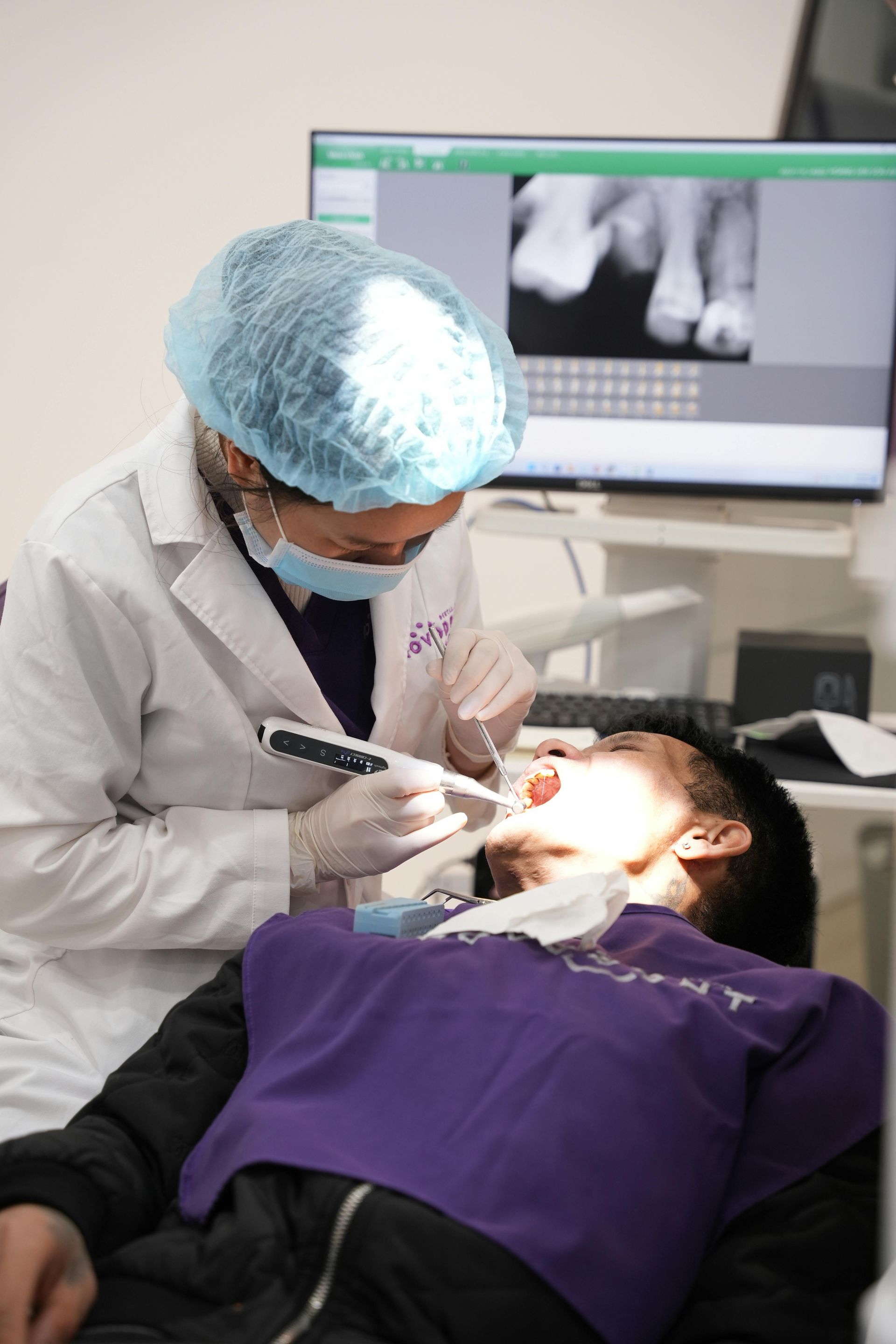 Dentist wearing mask and cap examines a patient's teeth. An X-ray is on the monitor in the background.