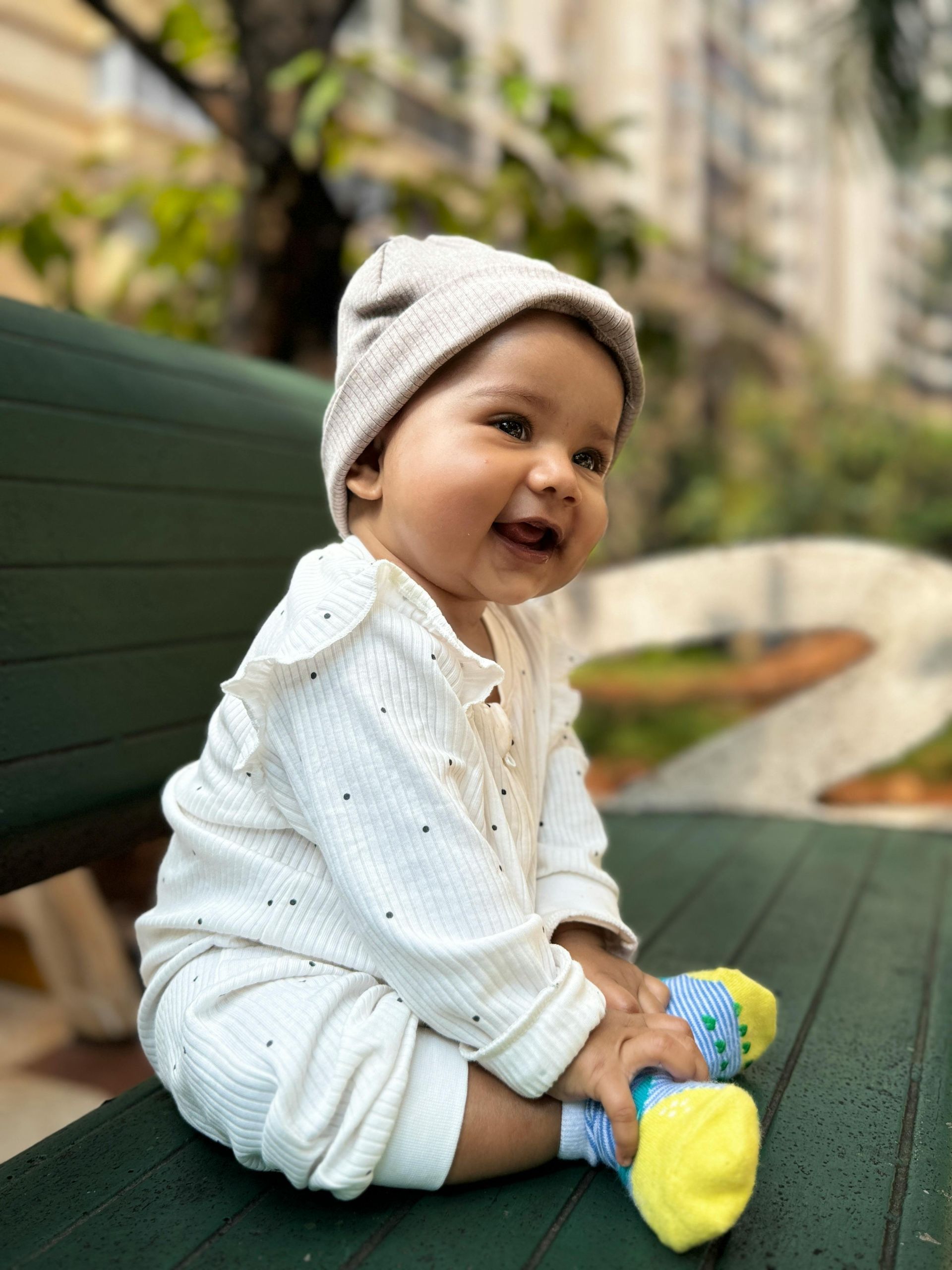 Smiling baby wearing a hat and white outfit, sitting on a park bench.